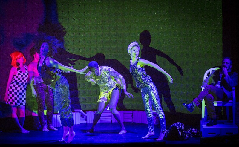 Two female dancers in sequin pans push their pelvises and arms behind them. Performers look on while Mark Dendy as Backhouse sits on a throne
